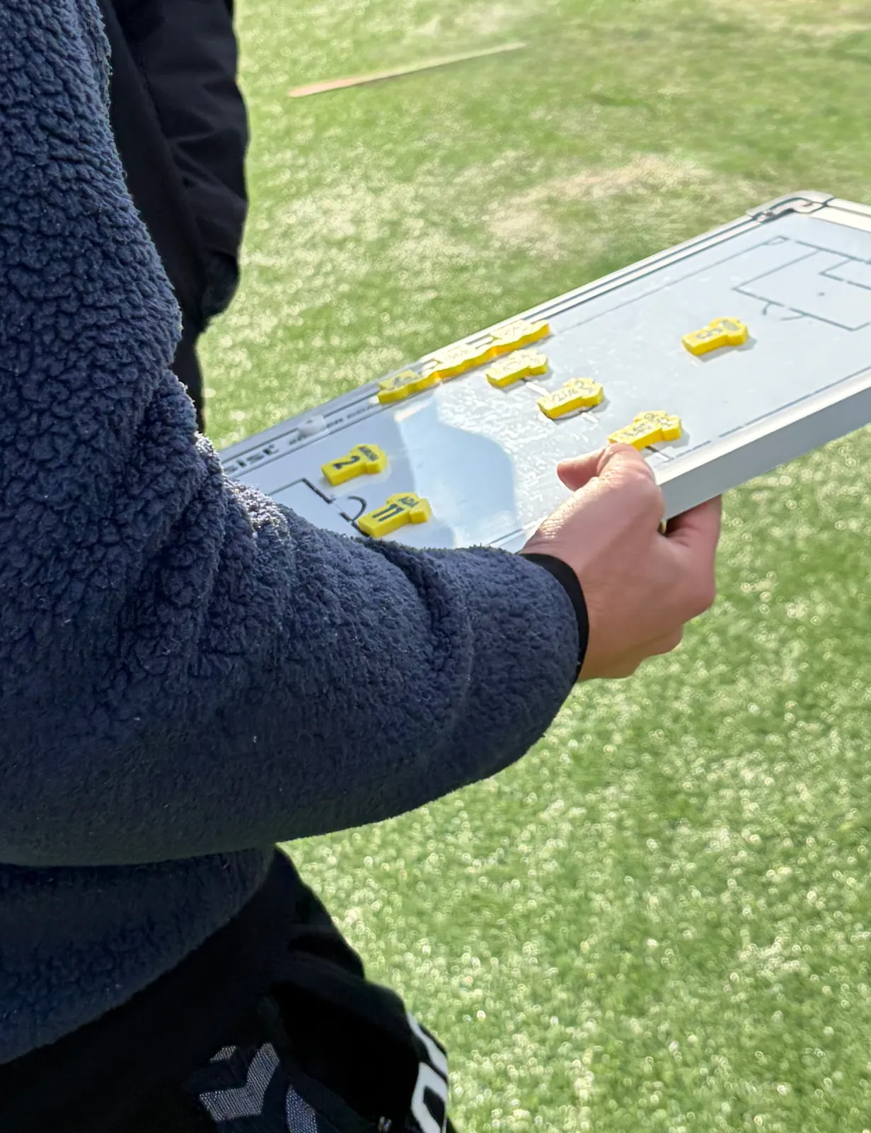Trener holder taktikktavla med draktmagneter i lagets farger ute på banen